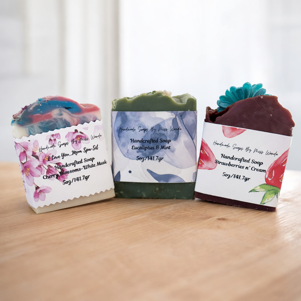 Three handmade soap bars displayed on a wooden surface. From left to right: 'I Love You More' soap with cherry blossoms and white musk, green eucalyptus and mint soap, and strawberries n' cream soap.